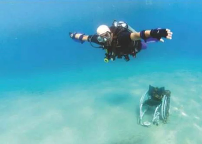 A scuba diver in a black wetsuit and gear swimming underwater with arms outstretched. A submerged object resembling a small boat is visible on the seabed below, surrounded by clear blue water.