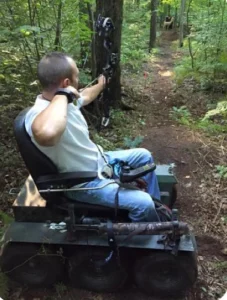 A person using a bow and arrow while seated on a specialized all-terrain wheelchair, positioned on a forest trail. The setting is surrounded by trees and greenery, highlighting wheelchair outdoor activities.