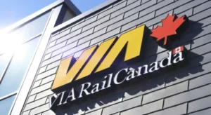 VIA Rail Canada logo displayed on a building's exterior, featuring bold yellow letters with a red maple leaf above it, against a gray wall. Sunlight reflects off the surface.