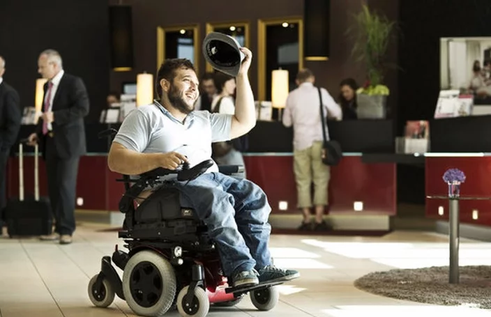 A man in a wheelchair wearing a cap, smiling and waving, in a modern hotel lobby. Other guests are visible in the background, engaging in conversations and checking in at the reception.