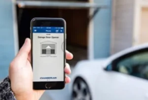A person holding a smartphone displaying a garage door opener app, with an open garage door visible in the background and a car parked nearby.