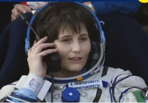 A female astronaut wearing a spacesuit and headset, communicating through a radio. She has short brown hair and is seated, with a focused expression, surrounded by various colors and people in the background.