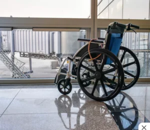 A wheelchair positioned near a large window at an airport, with reflections on a polished floor. In the background, a boarding bridge and terminal can be seen.