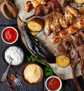 An assortment of grilled meats including chicken wings, skewers, and lamb, accompanied by roasted potatoes and dips in bowls on a wooden surface.