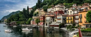 A picturesque lakeside town with colorful buildings along the water's edge, surrounded by lush greenery and mountains. Small boats are moored in the calm lake.