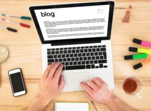 An overhead view of a person typing on a laptop with a document titled 'blog' open. Surrounding items include a smartphone, a notepad, colored highlighters, and a glass of tea, all placed on a wooden desk.