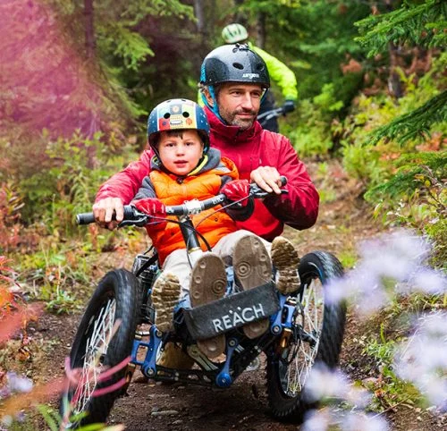A man with a kid wearing helmet & sitting in a wheelchair while greenery around