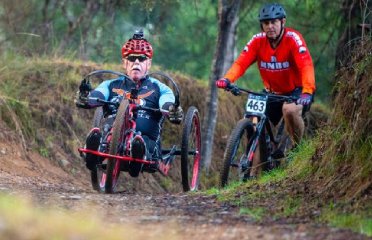 An athlete in a handcycle navigates a dirt path alongside another cyclist in a bright red jersey. Surrounding greenery and trees frame the outdoor scene.