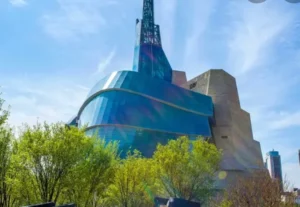 A modern, uniquely designed building with curved blue surfaces and angular concrete sections, surrounded by green trees and clear blue skies.