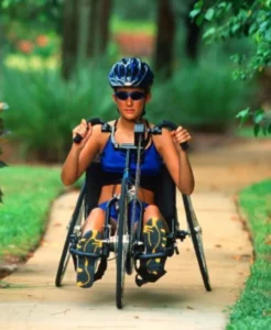 An athlete in a handcycle racing on a paved path, wearing a helmet and sunglasses, with greenery in the background.