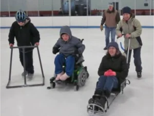 A group of individuals participating in adaptive ice skating on an indoor rink. Two children are using assistive devices for support while skating. One is in a wheelchair, and another is on a sled. The scene highlights accessible trails and fun activities for everyone.
