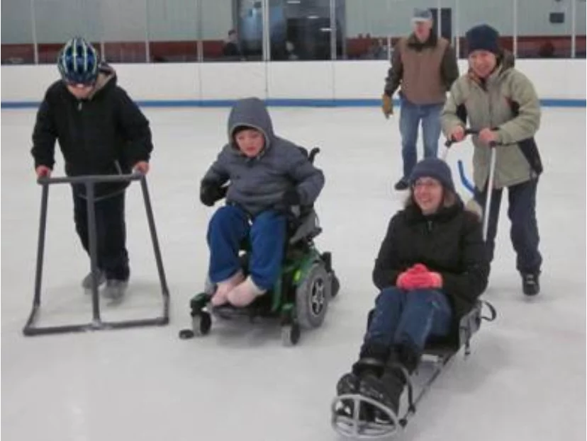 A group of individuals participating in adaptive ice skating on an indoor rink. Two children are using assistive devices for support while skating. One is in a wheelchair, and another is on a sled. The scene highlights accessible trails and fun activities for everyone.