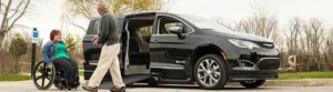 A person in a wheelchair smiles while talking to another person near a black wheelchair-accessible van with its sliding side door open in a parking lot.