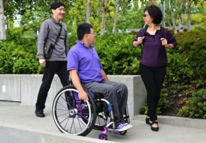 Three individuals are interacting outdoors in a park-like setting. One person is seated in a wheelchair, wearing a purple shirt, while two others are standing nearby in casual attire, conversing and smiling.