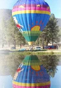 A colorful hot air balloon with a sun and star design is floating above a reflective pond, surrounded by trees and parked vehicles in a rural setting.