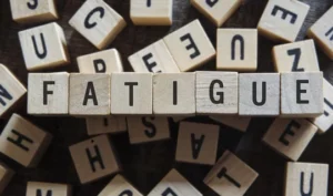 Wooden letter tiles scattered on a dark surface, spelling out the word 'FATIGUE'. The surrounding tiles include various other letters, emphasizing the central theme of fatigue in the context of Managing Spinal Cord Injury Fatigue.