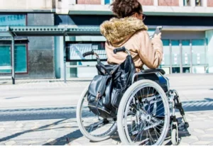A woman with curly hair seated in a wheelchair, wearing sunglasses and a tan jacket, using a smartphone on a city street. A black backpack is attached to the back of the wheelchair.