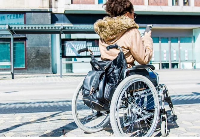 A woman with curly hair seated in a wheelchair, wearing sunglasses and a tan jacket, using a smartphone on a city street. A black backpack is attached to the back of the wheelchair.