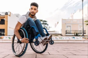A young man in a wheelchair smiles while posing outdoors. He is wearing a light t-shirt and jeans, with buildings and trees in the background.