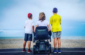 A woman sitting in a wheelchair & enjoying beach view while 2 kids standing around her