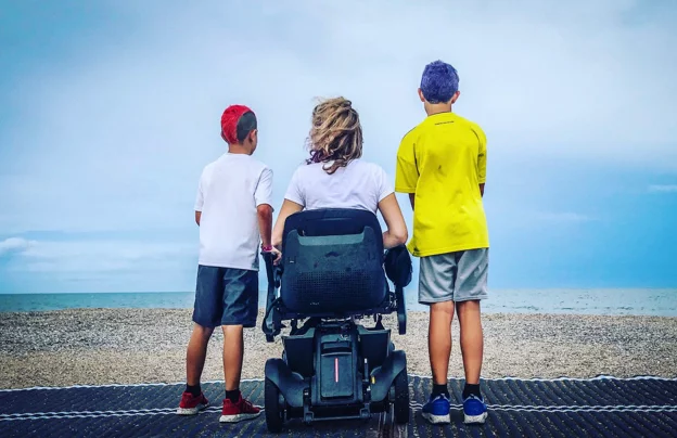 A woman sitting in a wheelchair & enjoying beach view while 2 kids standing around her
