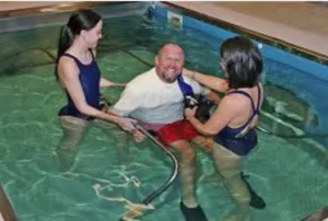 Two therapists assisting a man in a swimming pool, performing aqua therapy for spinal cord injury. The therapists are in swimwear, helping the man who is smiling and wearing a shirt, while supportive equipment is visible in the water.