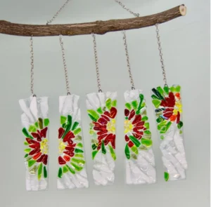 Four hanging glass panels featuring colorful floral designs in red, green, and yellow, suspended from a wooden branch by chains. The panels have a textured, crumpled appearance and are arranged in a row against a light background.