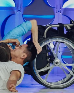 A performer in a wheelchair interacts closely with another performer on the stage, both engaged in a dynamic moment during a dance performance.