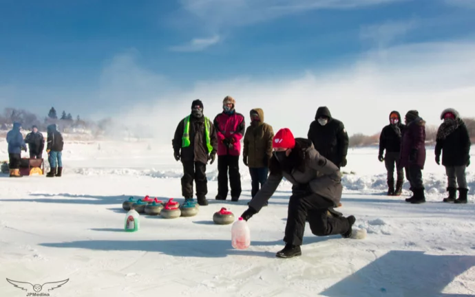 A wheelchair curling event on an icy surface, with participants dressed warmly and some spectators in the background. A player is preparing to release a curling stone, while others observe. Snowy landscape under a clear sky.
