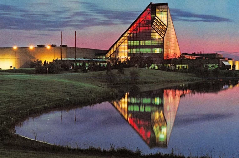 A modern glass building with a triangular roof reflecting in a calm pond at dusk, surrounded by greenery and softly lit by exterior lighting.