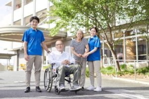 A male caregiver in blue shirt assisting an elderly man in a wheelchair, alongside a middle-aged woman and a female caregiver in a blue shirt. They stand outside a healthcare facility with greenery in the background, depicting a supportive environment, emphasizing Indeed healthcare jobs.