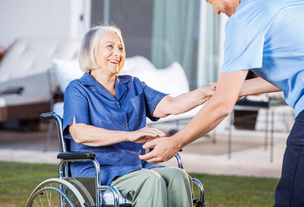 An elderly woman sitting in a wheelchair smiles as a caregiver reaches out to assist her. They are outdoors with a blurred background of patio furniture, offering a scene of support and mobility assistance.
