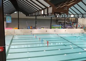 An indoor swimming pool with clear blue water and lane dividers. Colorful pool floats and slides are visible in the background. The facility features a high ceiling with wooden beams and overhead lights.