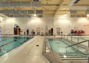 An indoor swimming pool area at YMCA West Winnipeg. The image shows a large swimming pool on the left and a smaller accessible pool on the right, with lifeguards and equipment present.