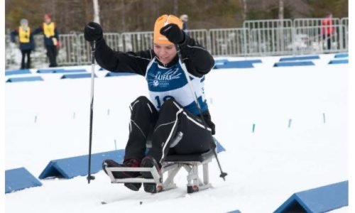 An athlete in a competitive sled racing event, wearing a yellow helmet and black gear, is pushing forward on a snowy track while seated on a sled. The background features barriers and supporters. This event promotes Accessible Sport Connection in Winnipeg, Manitoba.