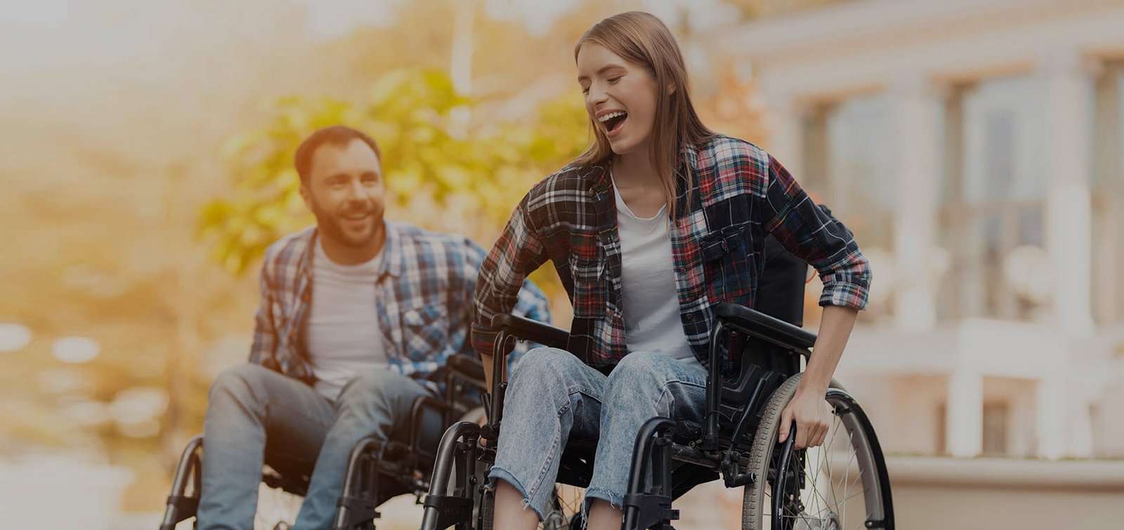 Two individuals in wheelchairs enjoying a sunny day outdoors, smiling and engaging with each other. They are dressed casually in plaid shirts and jeans, with a blurred background of green trees.