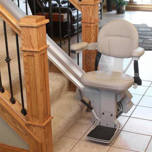 A stairlift positioned at the bottom of a staircase, featuring a padded seat and armrests, with a metal track installed along the stairs. The home interior includes light-colored walls and a tiled floor.