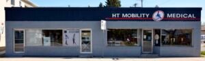 An exterior view of the HT Mobility Medical storefront, featuring a sign with the business name prominently displayed. The building has large windows, and a poster with an illustration of a person is visible on the left side.