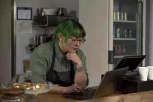 A person with green hair wearing glasses and an apron is working on a laptop at a café counter. They are focused on the screen, with some pastries displayed in front of them, suggesting a busy work environment in a café.