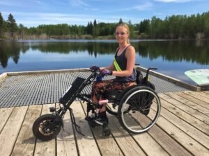 Young lady is sitting on a dock by wilderness Lake with your adaptive wheelchair, custom equipment, and wearing adaptive clothing made for the outdoors