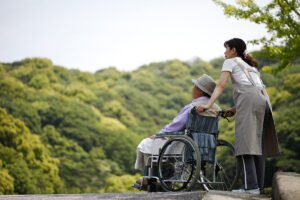 Healthcare aid pushing an elderly man in a wheelchair on a path near some hills and trees
