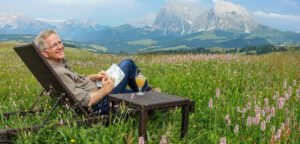 Beautiful landscape, photo of Rick Steve’s lounging in a nice easy big chair on an accessible green field with majestic snow, cap mountain peaks in the background and a field full of flowers