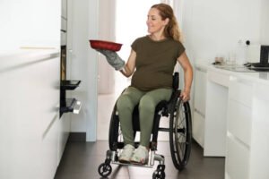 A woman wearing brown shirt and green pants in her wheelchair is in her accessible kitchen, taking a dish out of the oven and you can also see the cabinets on the other side have allowance for her to roll under the counter in order to work easier