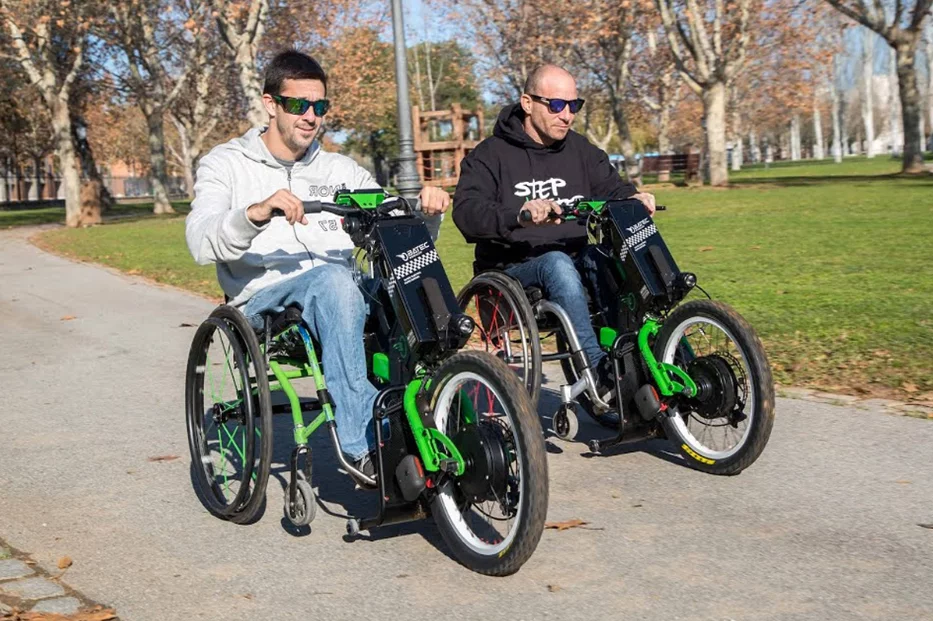 2 men wearing glasses & sitting in wheelchairs while greenery & tress around