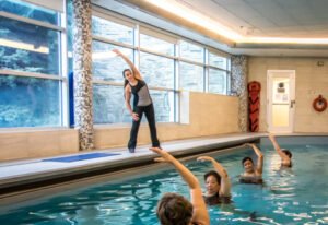 A fitness instructor leading an exercise class in a swimming pool. Participants are in the water, following the instructor's movements with raised arms, in a well-lit indoor setting with large windows.