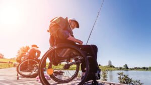 Two individuals in wheelchairs fishing from a wooden dock by a peaceful lake. The setting is sunny, with clear blue skies and greenery in the background, highlighting accessible outdoor attractions in Manitoba.