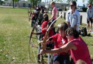 A group of individuals in wheelchairs practicing archery in a grassy field. Instructors assist participants, highlighting the Accessible Sport Connection Manitoba initiative.