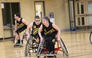 Three wheelchair basketball players in team uniforms, focused on the game in a gymnasium. One player is leading while the others follow closely, all engaged in the sport. The scene highlights Inclusive Recreation in Manitoba.