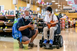 A man in a blue shirt and shorts crouches next to a woman in a mobility wheelchair inside a grocery store, both wearing masks. The store has shelves filled with various products in the background, emphasizing accessibility.