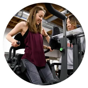 Two women interacting at a gym. One woman is using a fitness machine while the other is providing assistance or guidance. Both are focused on the exercise in a well-lit indoor fitness facility.